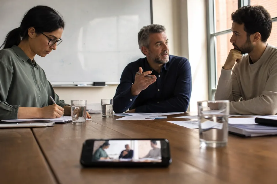 Three colleagues around a meeting table, a phone recording in the middle, one of them taking notes